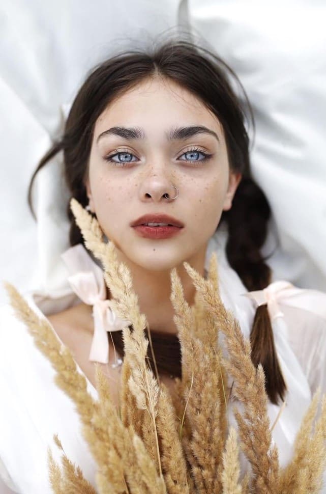 Woman with braided hair and pink ribbons lies on white fabric in a white blouse, holding dried wheat stalks.