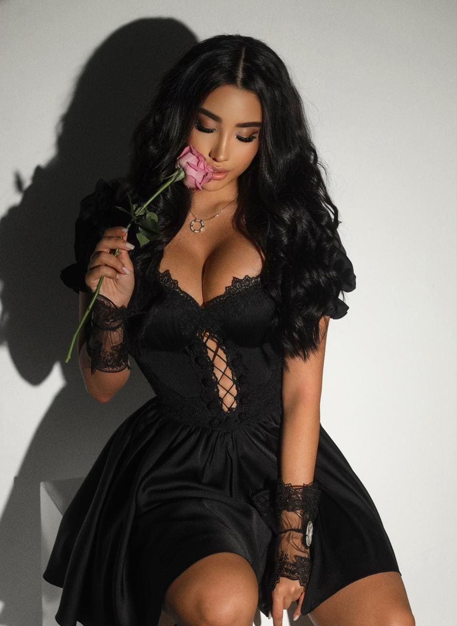 A seated woman in a short black lace-trimmed dress holds a pink rose near her face against a plain light wall.