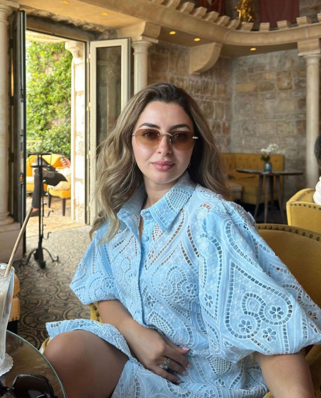 A woman in a light blue eyelet dress and sunglasses sits in a yellow chair near a garden doorway.