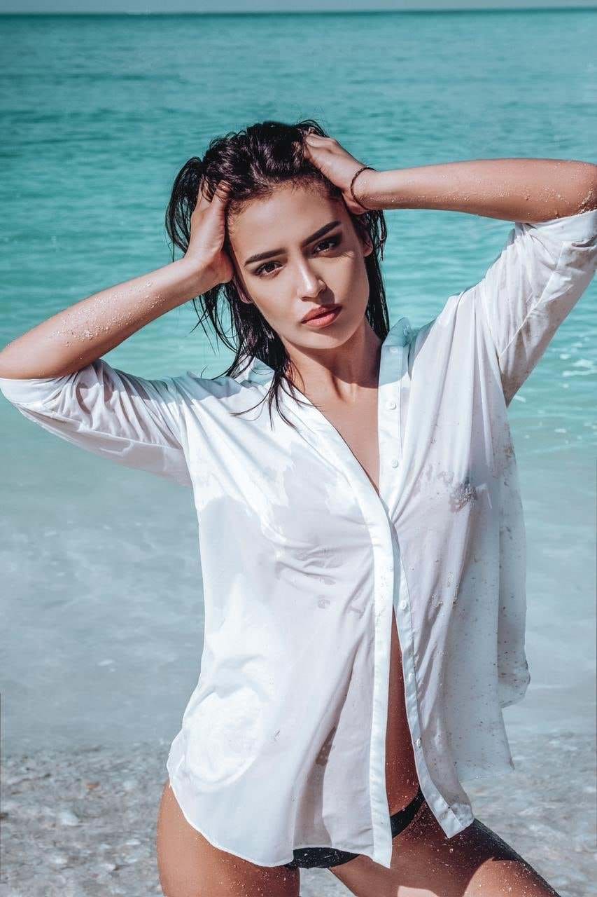 A woman with wet hair stands on a beach in an unbuttoned white shirt and black bikini bottom, holding her head.
