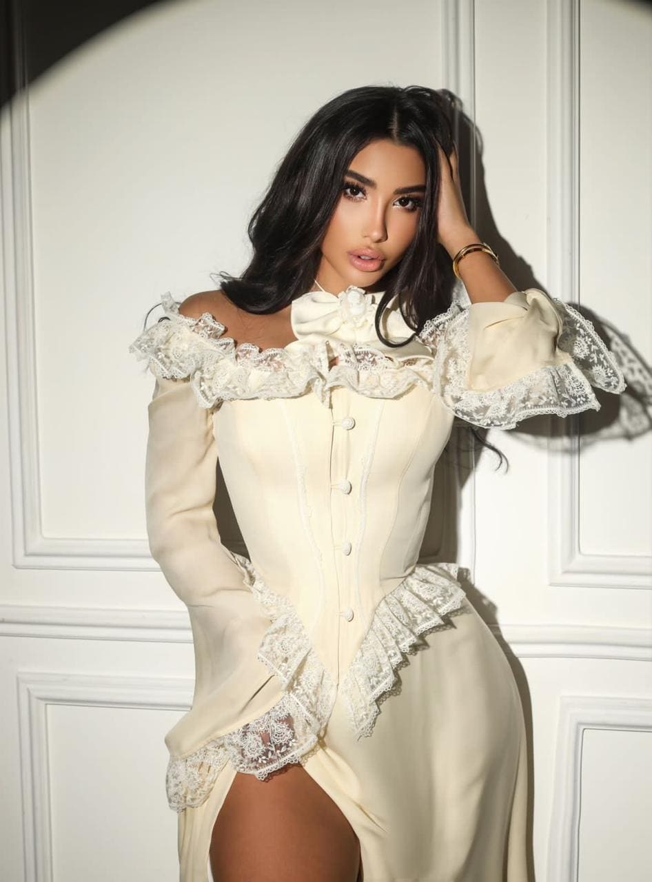 A woman in a cream off-the-shoulder lace dress with a high slit stands against a white paneled wall, hand raised to her head.