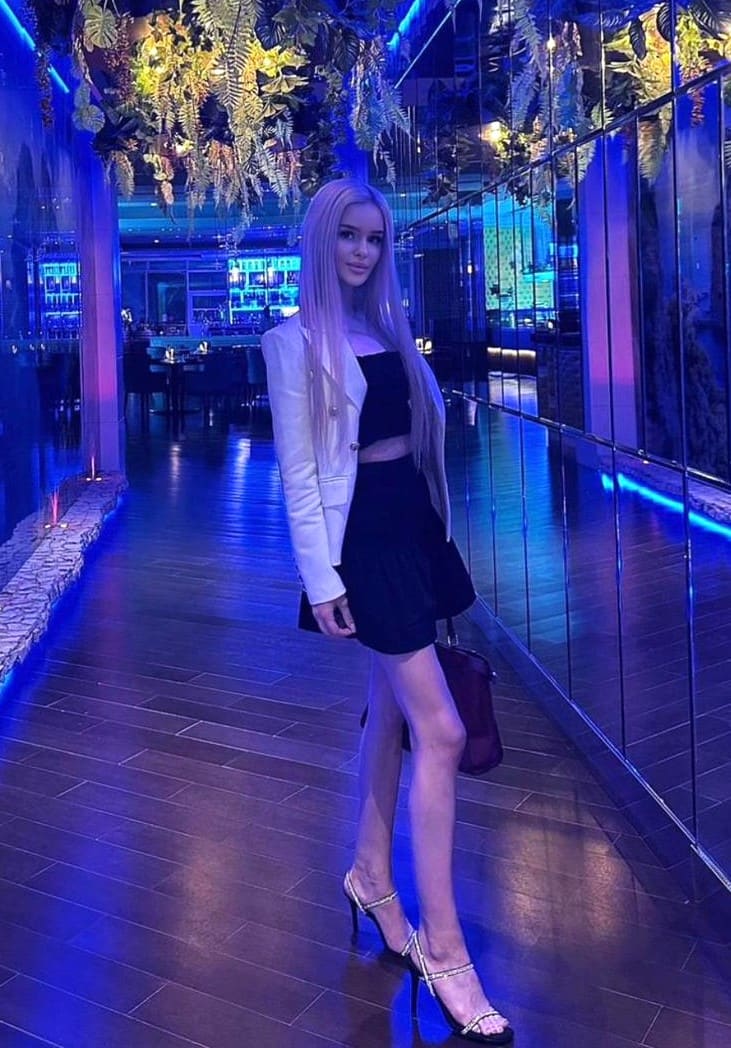A woman in a white blazer, black crop top, and short skirt stands in a dim corridor with blue lighting and plants.