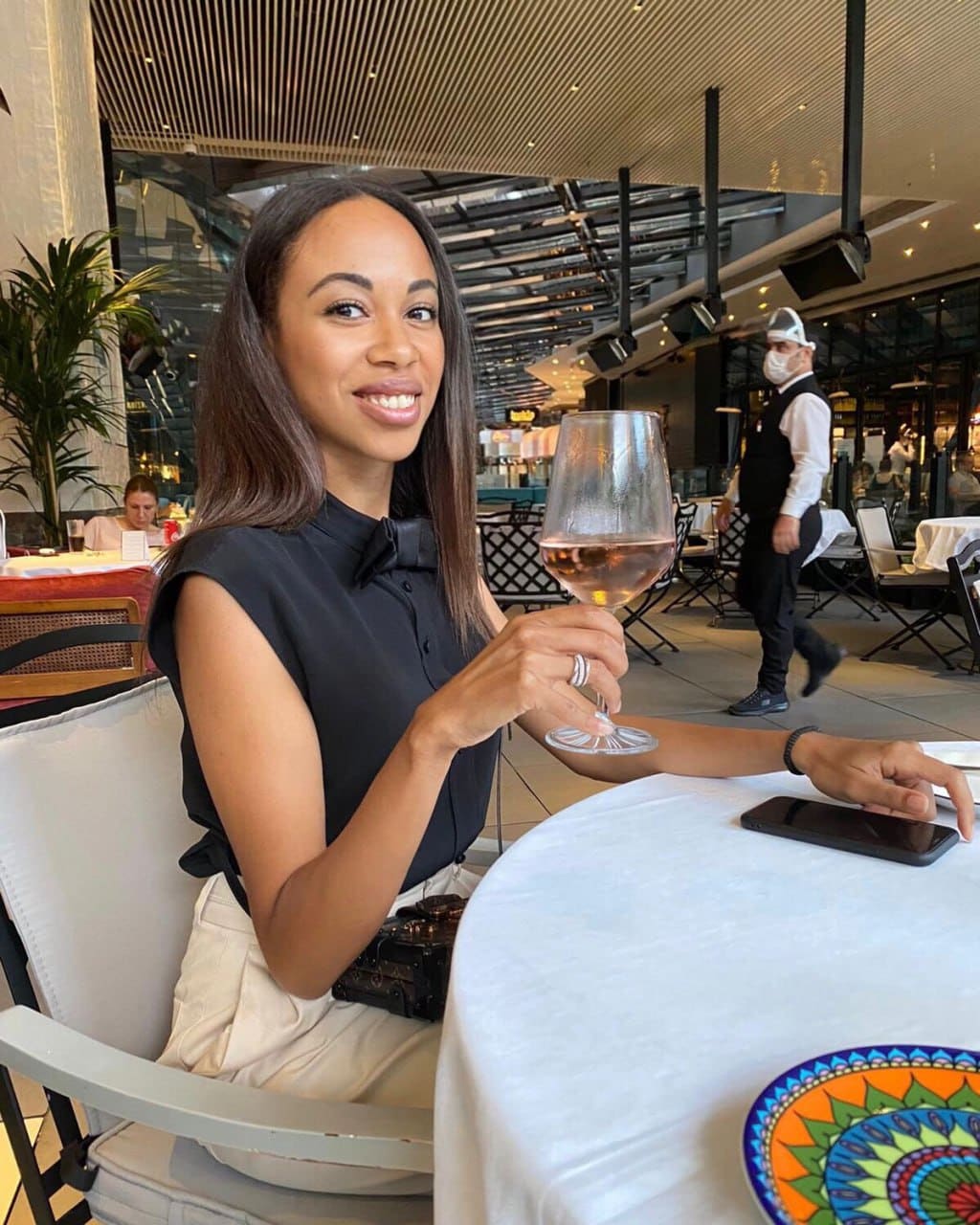 A woman sits at a restaurant table holding a glass of rosé wine, wearing a black sleeveless top and light trousers, with a phone nearby.