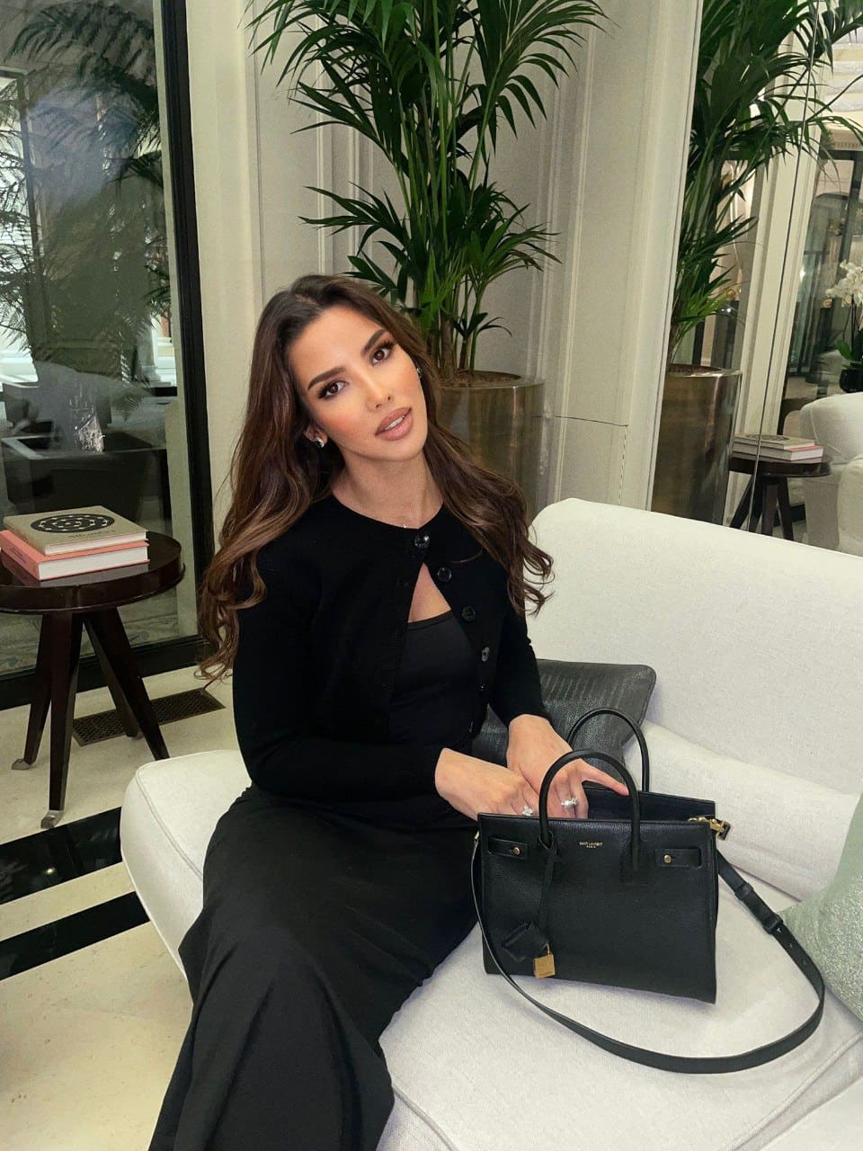 Louisa sits on a white sofa in a black dress with a keyhole neckline, holding a handbag, surrounded by plants and books.