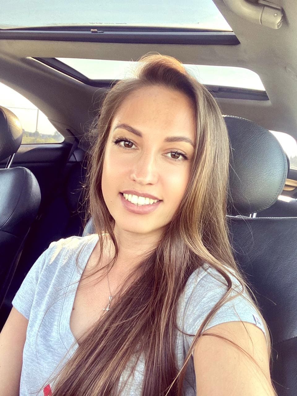 A woman smiles from a car driver's seat with a sunroof, wearing a grey t-shirt and necklace, with long brown hair.