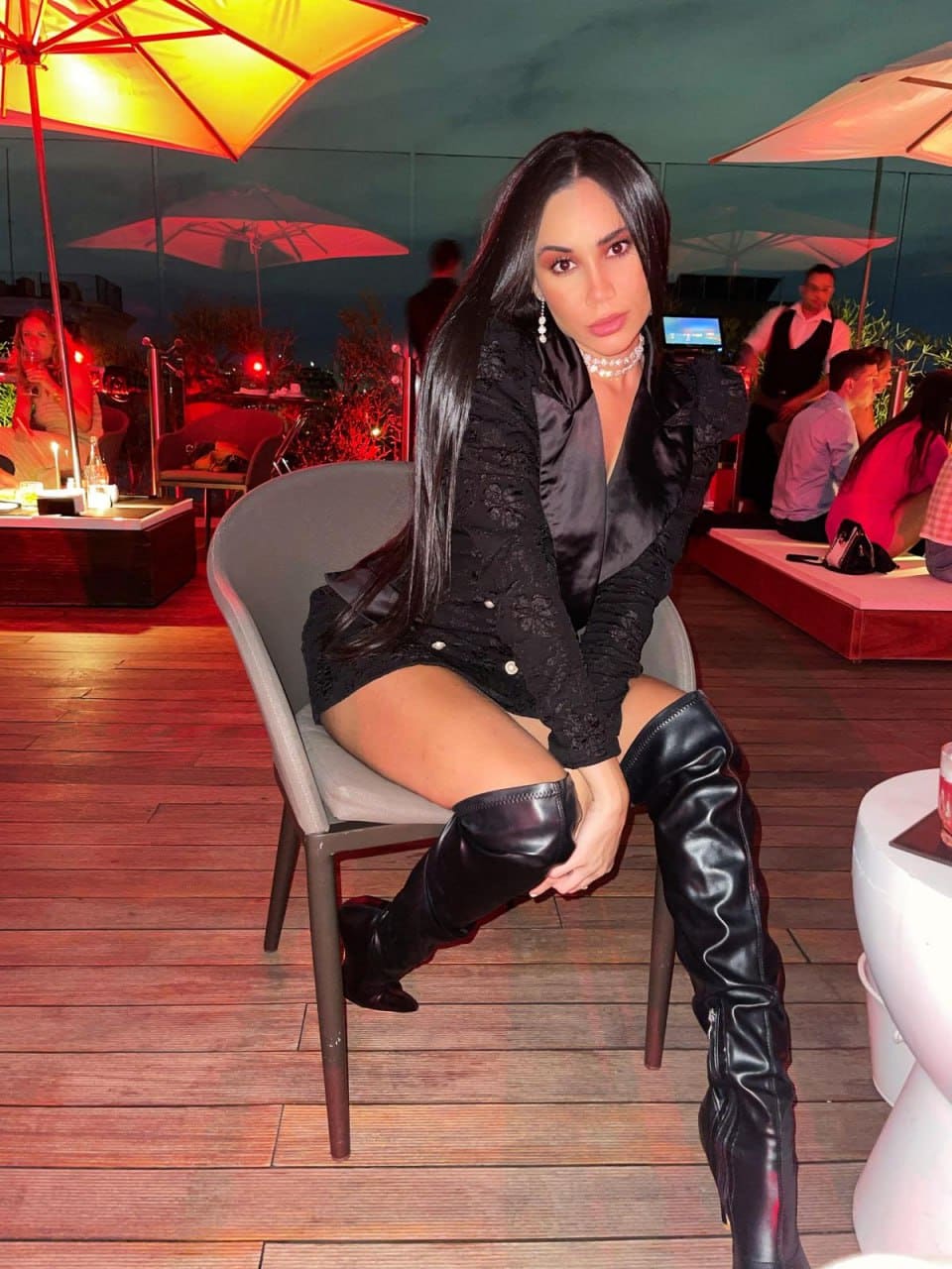 She sits on a grey chair at an outdoor evening venue, wearing a black lace jacket, dark satin top, short skirt, and thigh-high boots.