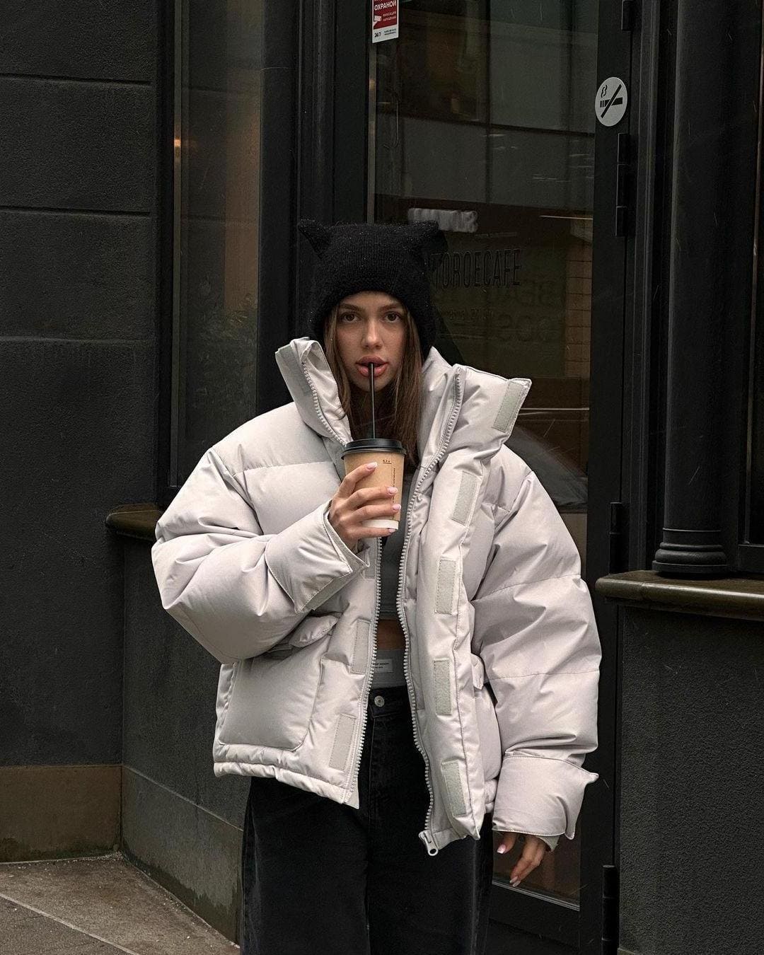 A woman in a cat-ear beanie and grey puffer jacket sips coffee from a straw on a city sidewalk outside a dark storefront.
