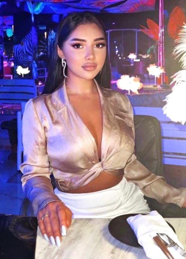 Seated woman in a beige V-neck blouse and white skirt rests a hand on a table in a dimly lit venue.