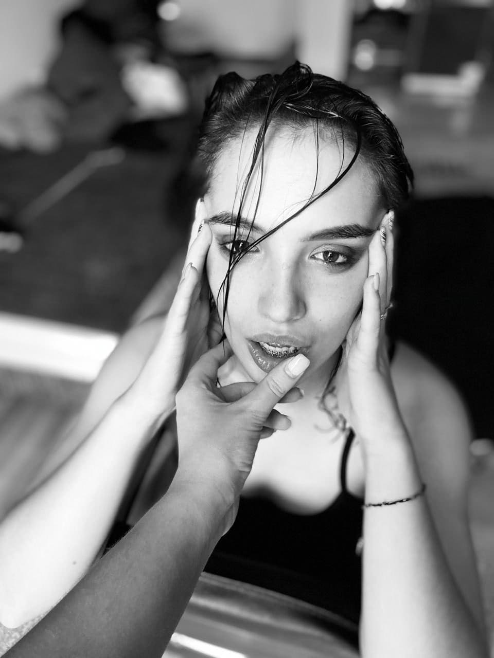 Black-and-white portrait of a woman with slicked-back hair and dark makeup in a black tank top, hands framing her face.