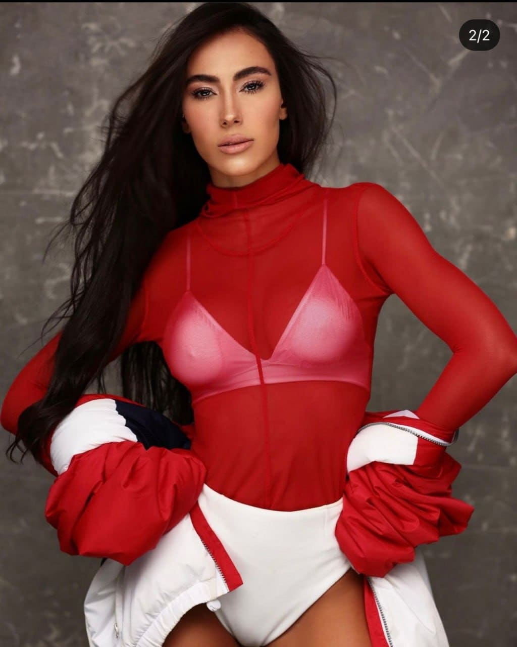 A woman stands hands on hips in a sheer red top, pink bra, white bottoms, and a red and white jacket draped over her arms.
