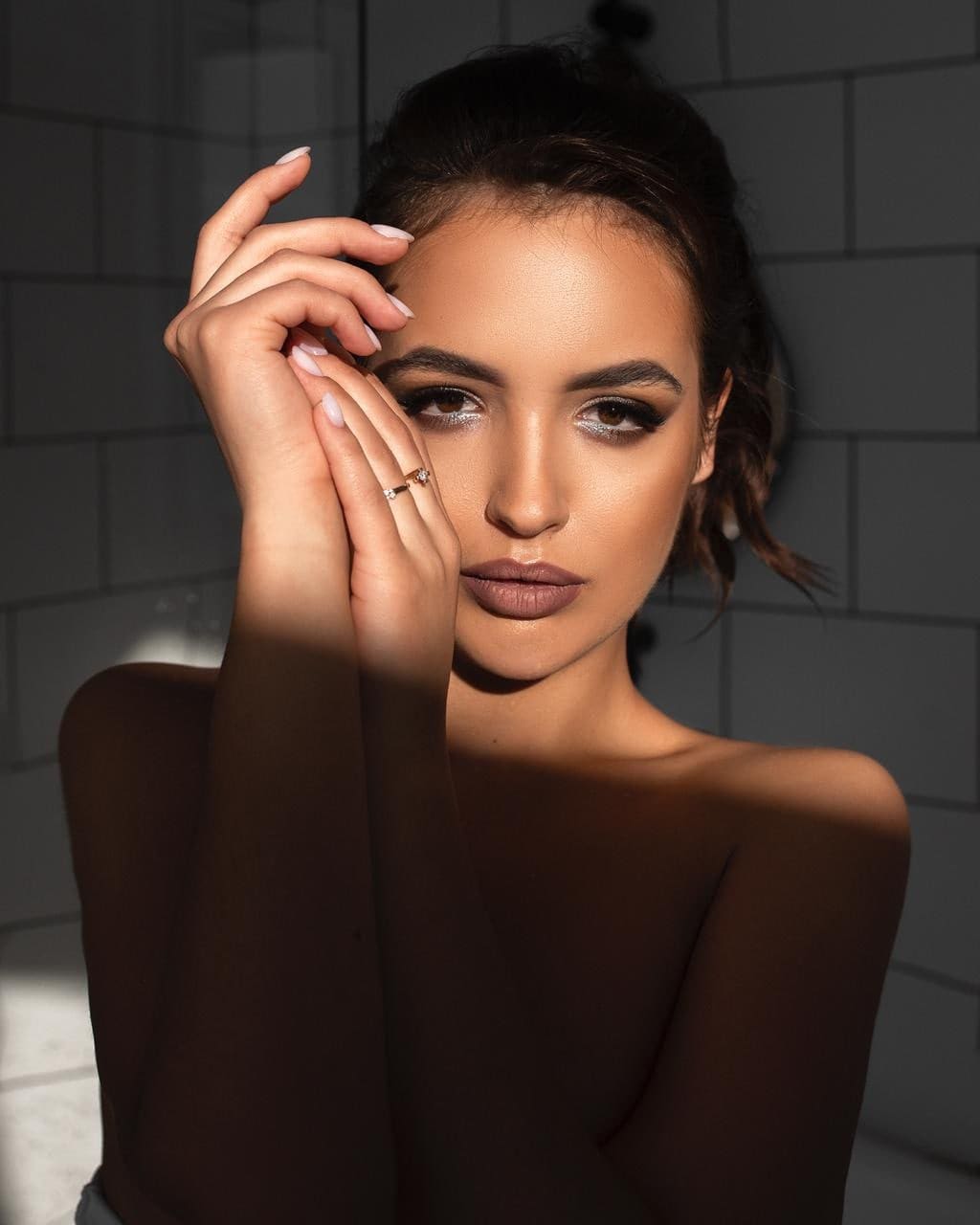 Uliana poses with hands near her face, wearing a dark off-the-shoulder top against a tiled wall.