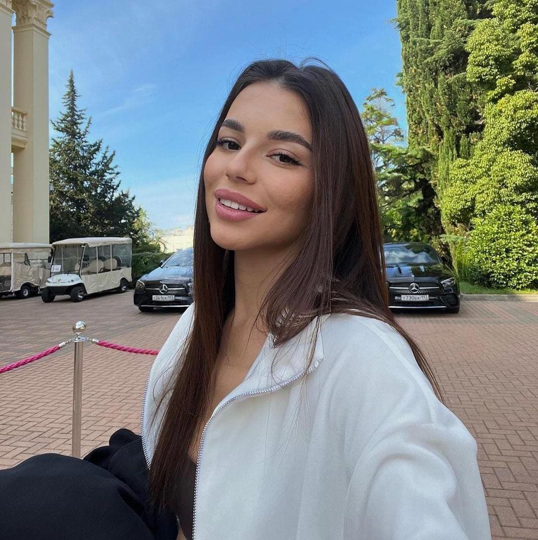 Annabelle takes a selfie outdoors in a white zip-up jacket over a dark top, with cars and trees behind her.