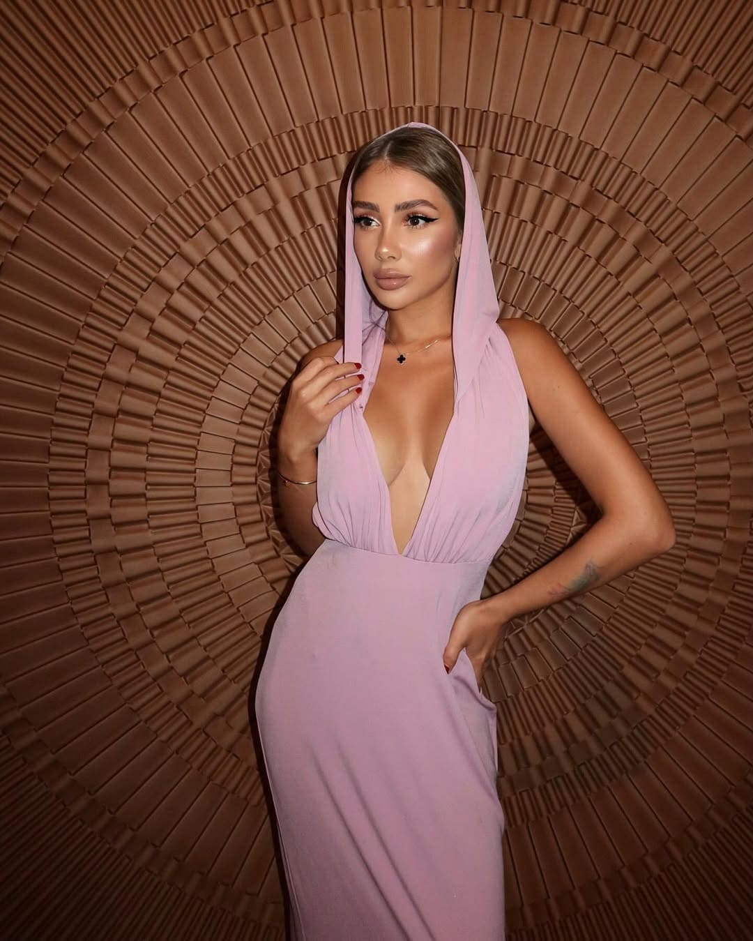 A woman in a light pink hooded dress with a deep V-neck stands before a brown pleated fan backdrop, one hand on hip.