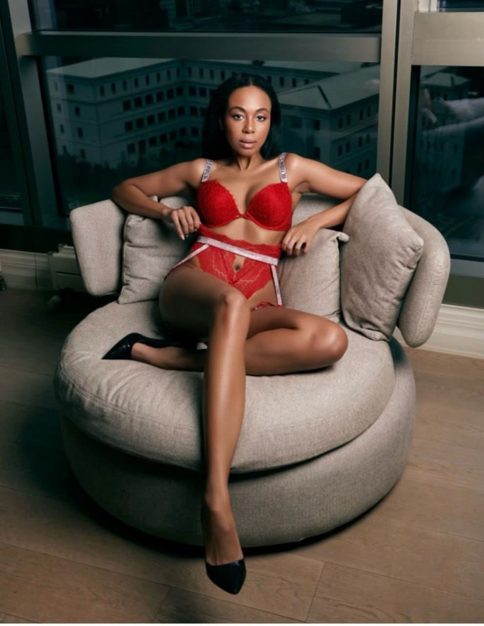 A woman sits on a beige armchair wearing a red lace bra and briefs with white trim, paired with black pointed-toe heels, near large windows.
