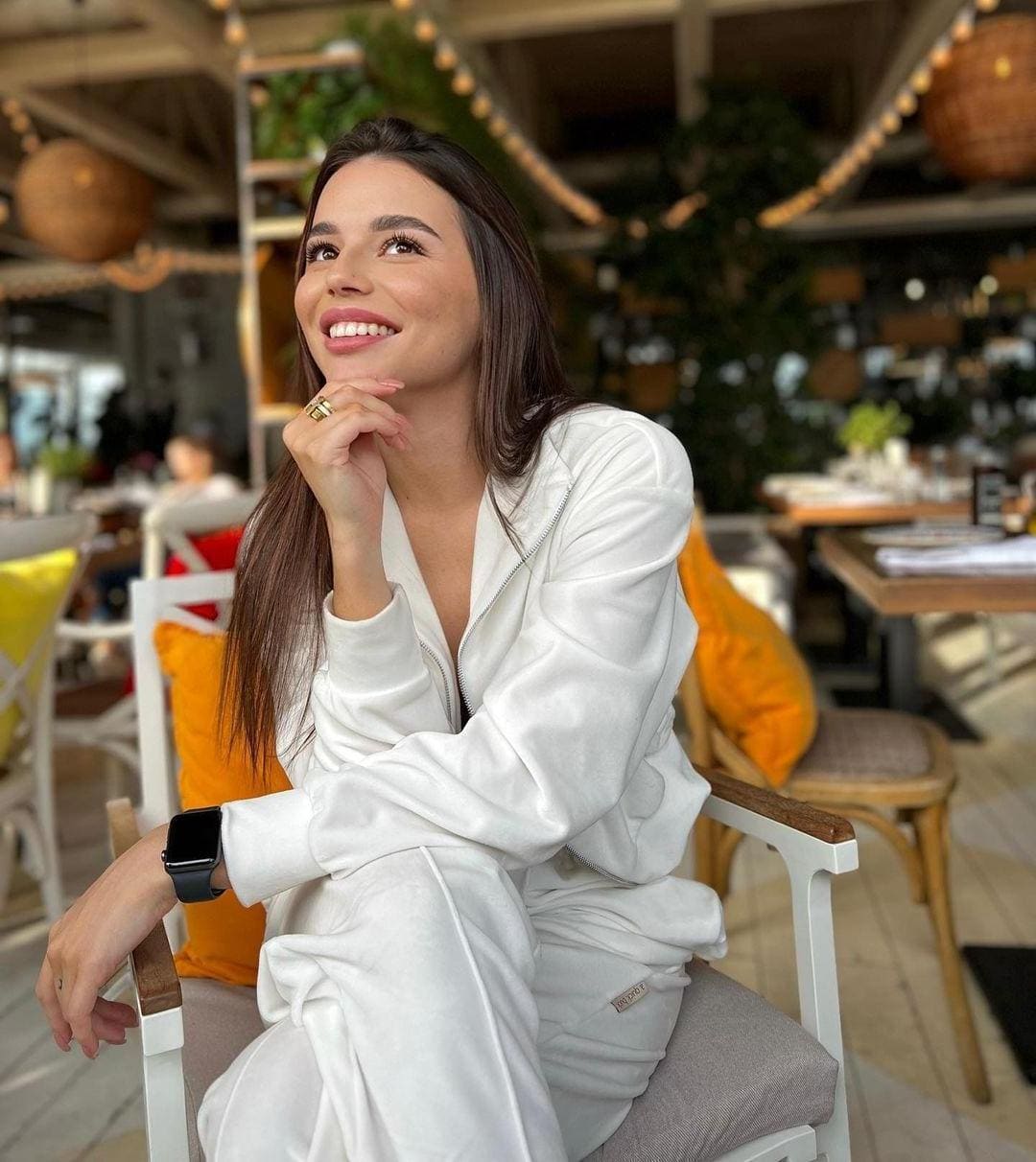 A woman sits in a white chair at a cafe wearing an open white tracksuit, resting her chin on her hand.