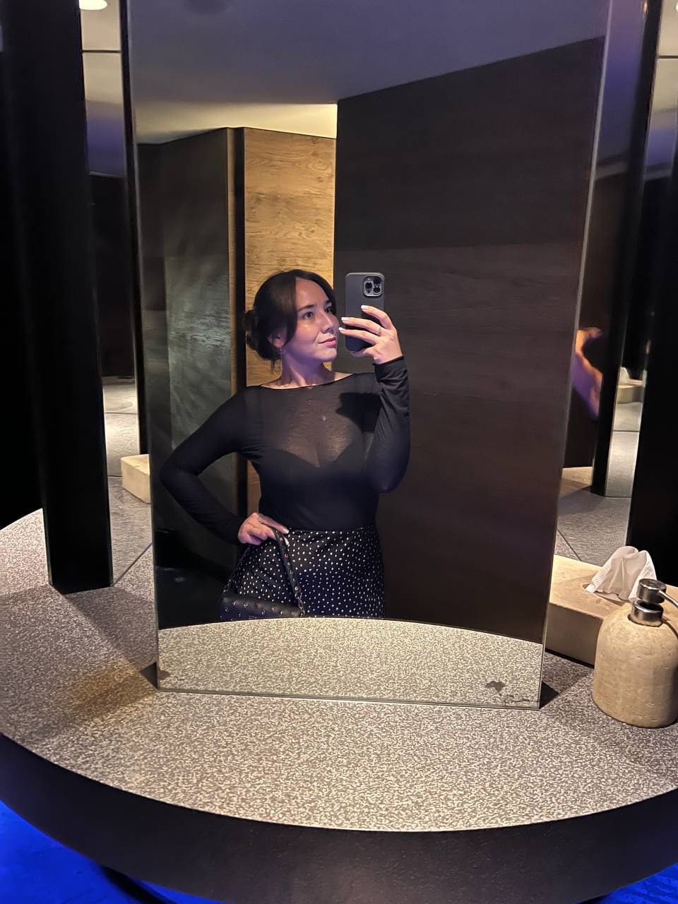 Woman in a sheer black top and polka-dot skirt takes a mirror selfie with one hand on her hip.