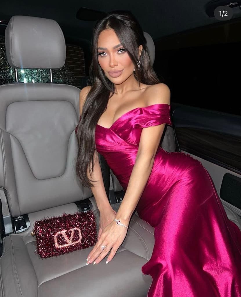 In a car, she wears an off-the-shoulder magenta satin gown with a clutch beside her.