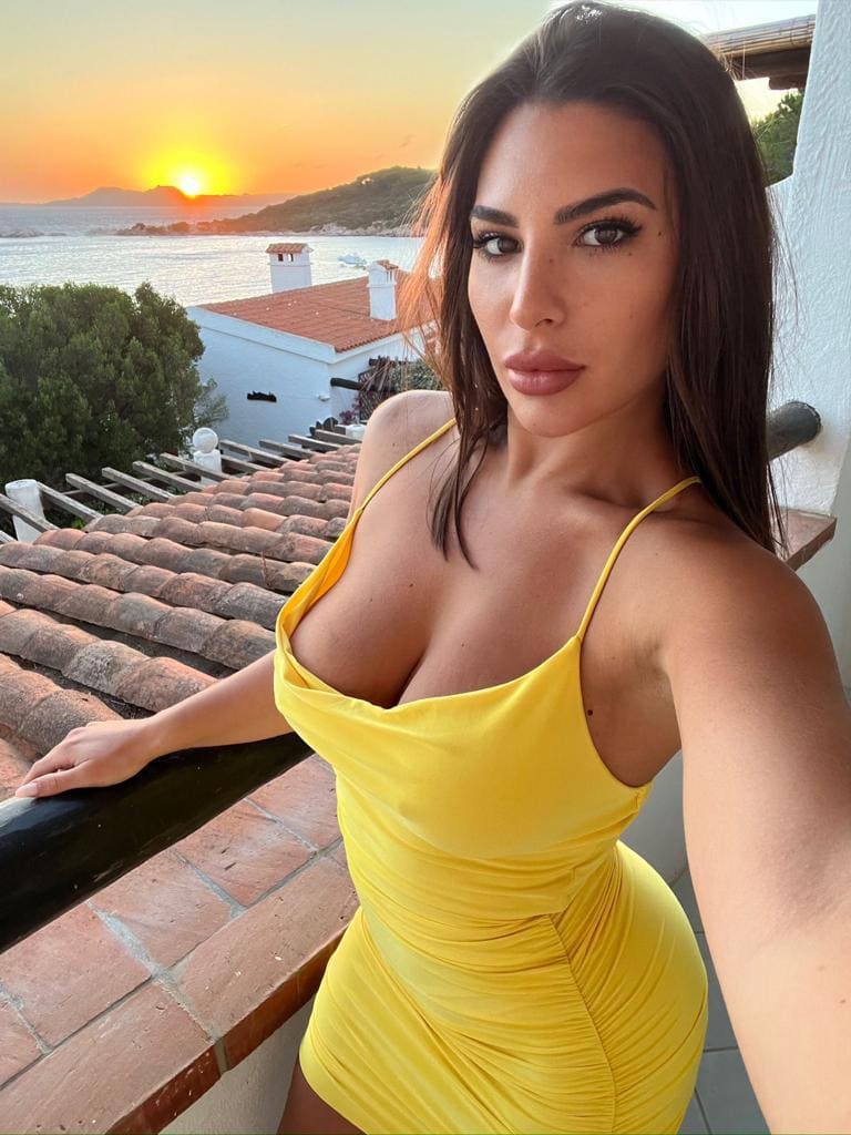 Rebecca stands on a tiled balcony at sunset in a short yellow dress, resting her hand on the brick railing with the sea behind.