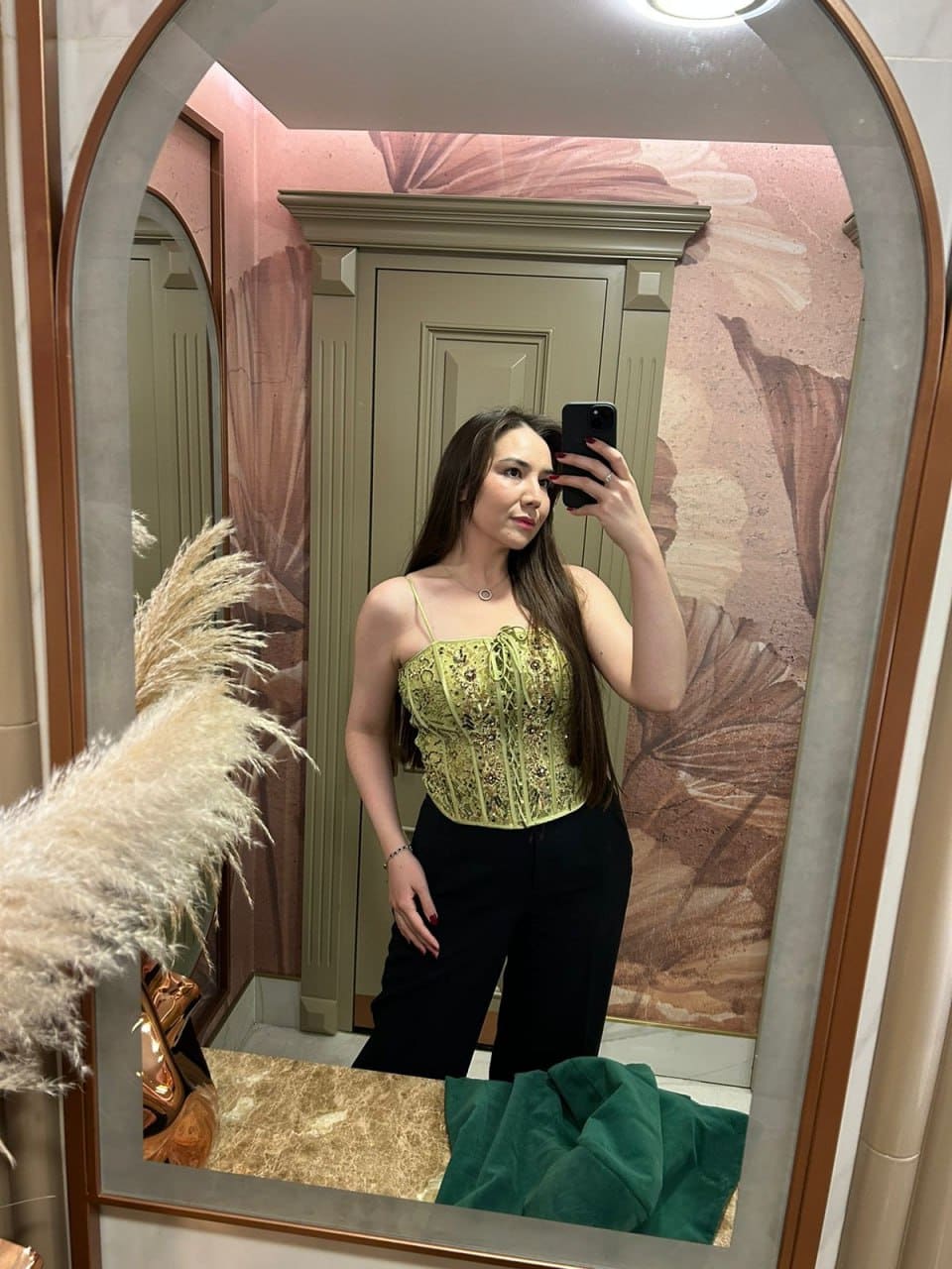 Bathroom mirror selfie featuring a woman in a green lace camisole and black trousers near floral wallpaper.