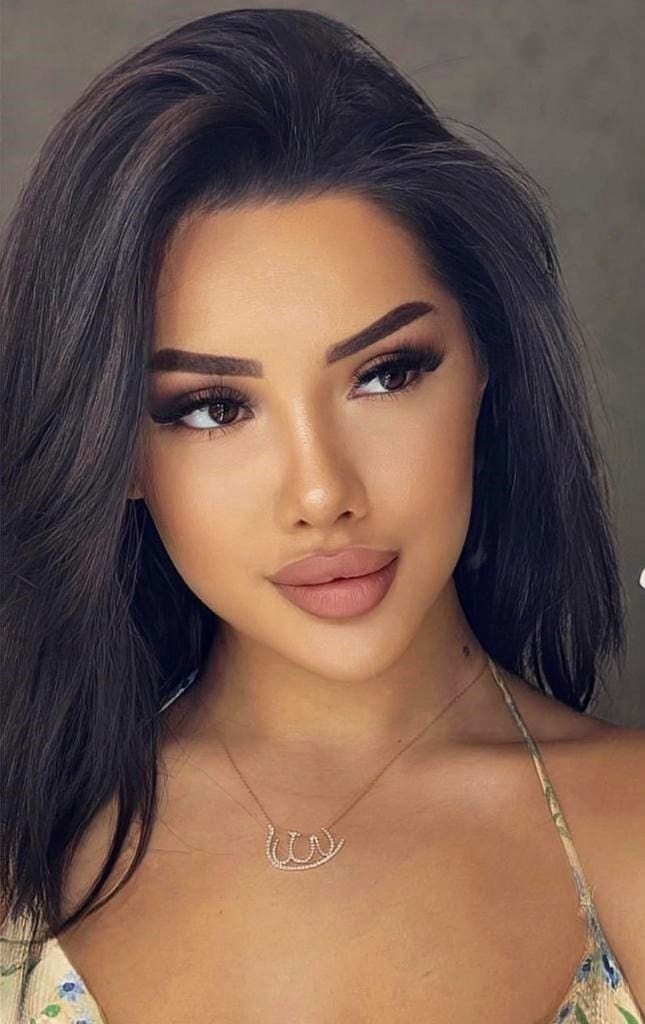 Valentina with long dark hair and heavy eye makeup wears a floral top with thin straps and a necklace against a gray background.