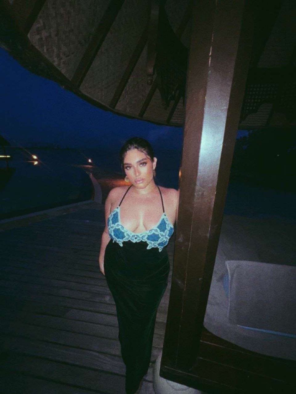 Leaning against a post on a night deck, she wears a long black dress with a blue lace halter neckline, set against dark water.