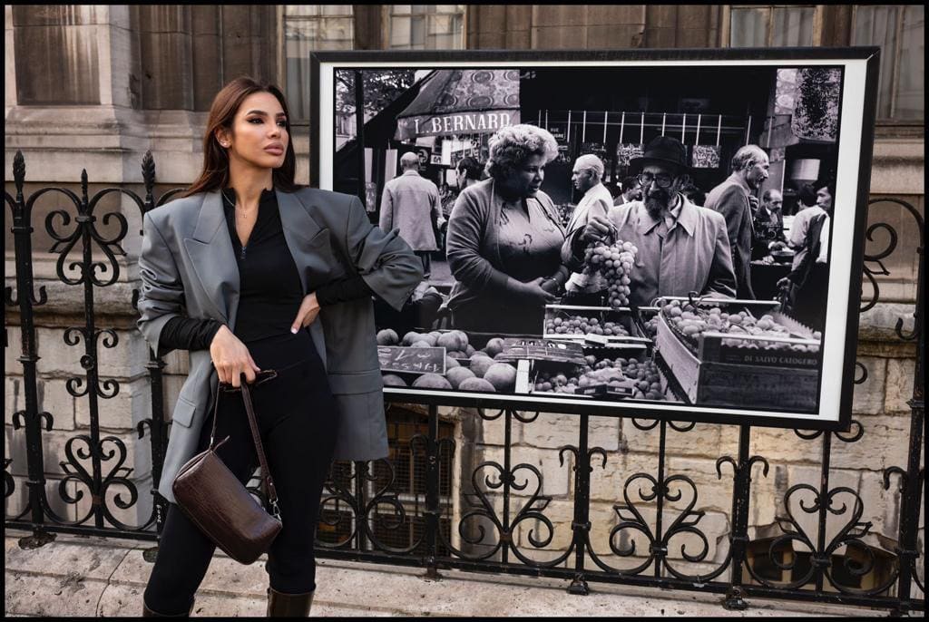 A woman in a grey blazer and black pants stands before a framed photo, holding a brown bag, with an iron fence behind.