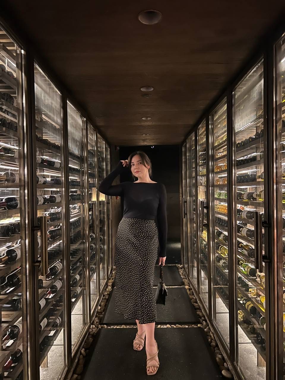 Woman in a black top and polka-dot skirt stands in a glass wine cellar corridor, touching her hair.