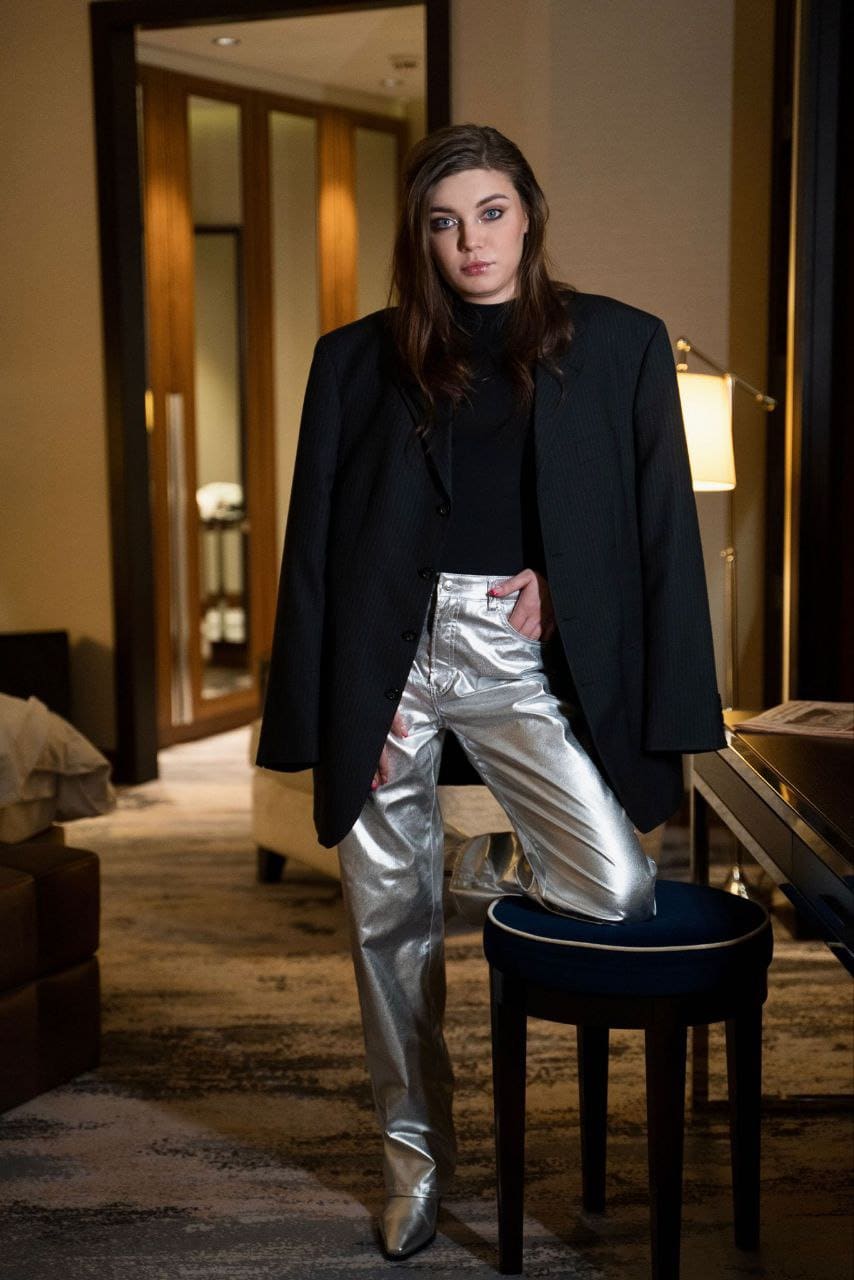 Lilly stands in a hotel room wearing a black turtleneck, oversized blazer, and silver trousers, with one foot on a blue stool.