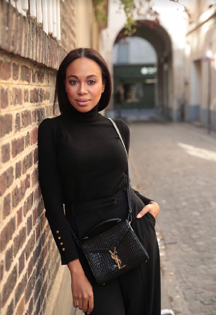 Destina stands against a brick wall in a narrow street, wearing a black turtleneck, black trousers, and a textured crossbody bag.