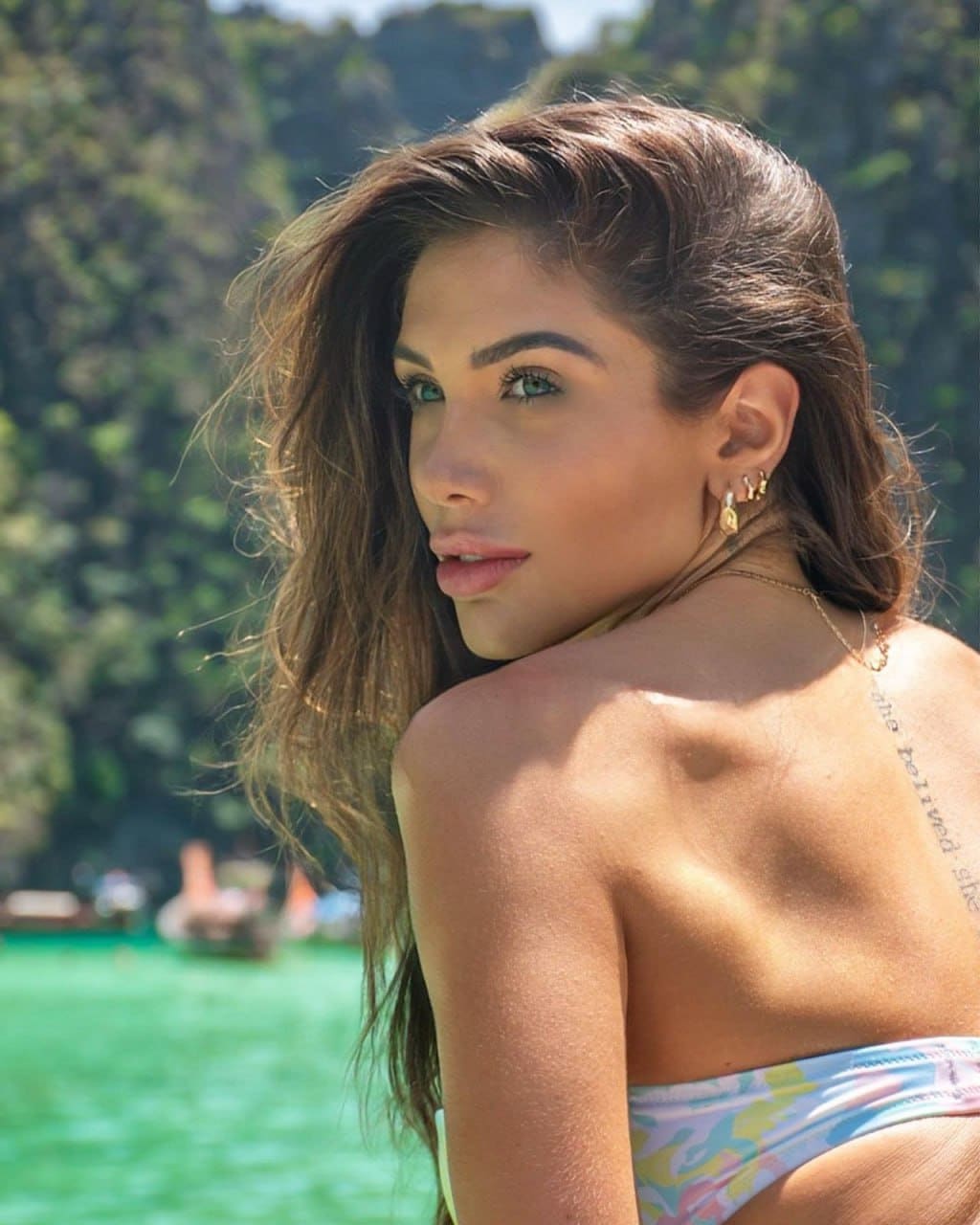 Seen from behind, a woman in a patterned bikini top looks over her shoulder against a green lake and forested hill.