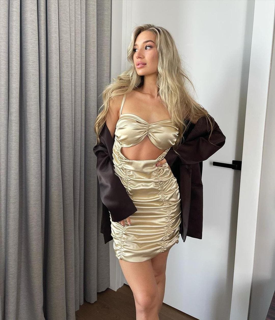 A blonde woman stands in a room with grey curtains, wearing a champagne satin dress with a midriff cutout and a brown blazer over her shoulders.