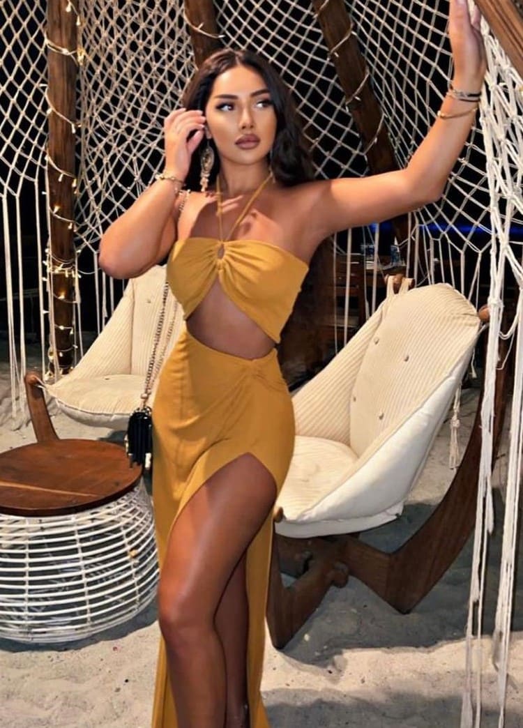 Woman in a mustard-yellow cut-out dress poses with a raised hand against rope netting and white chairs.