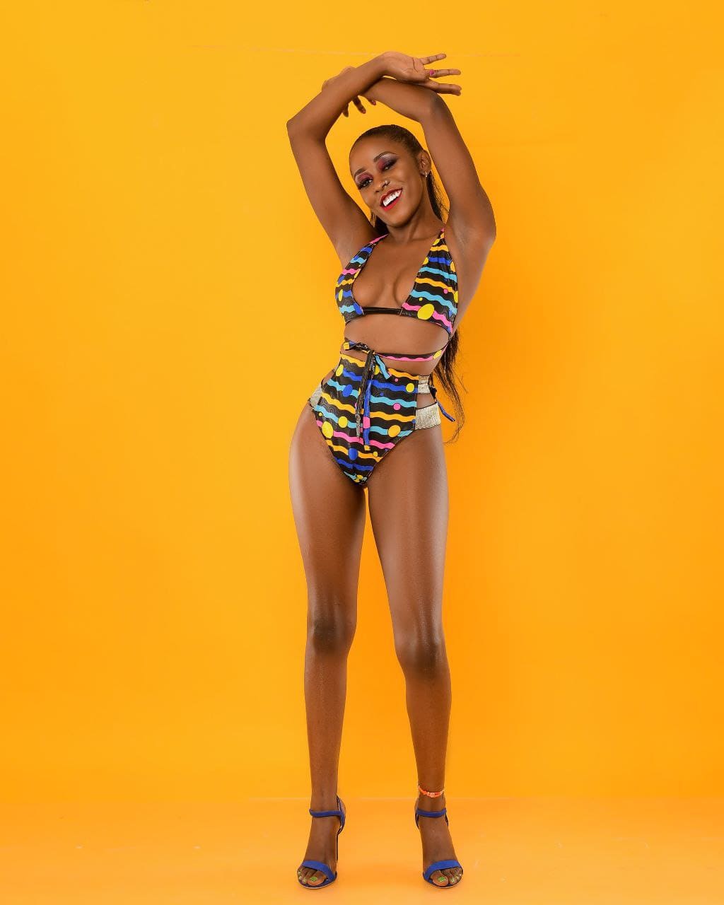 A woman in a patterned halter swimsuit and blue heels stands against yellow with her arms raised above her head.