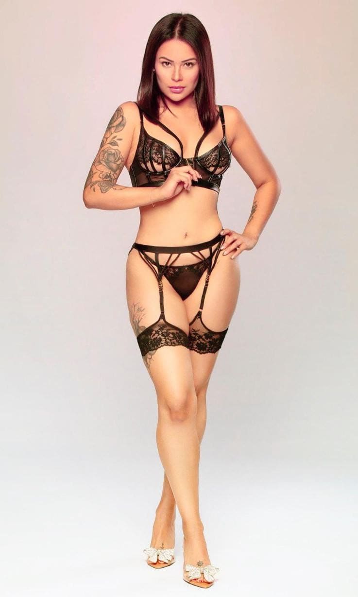 A woman with tattoos stands in a studio wearing black lace lingerie with a garter belt, stockings, and white heels, hand on hip.