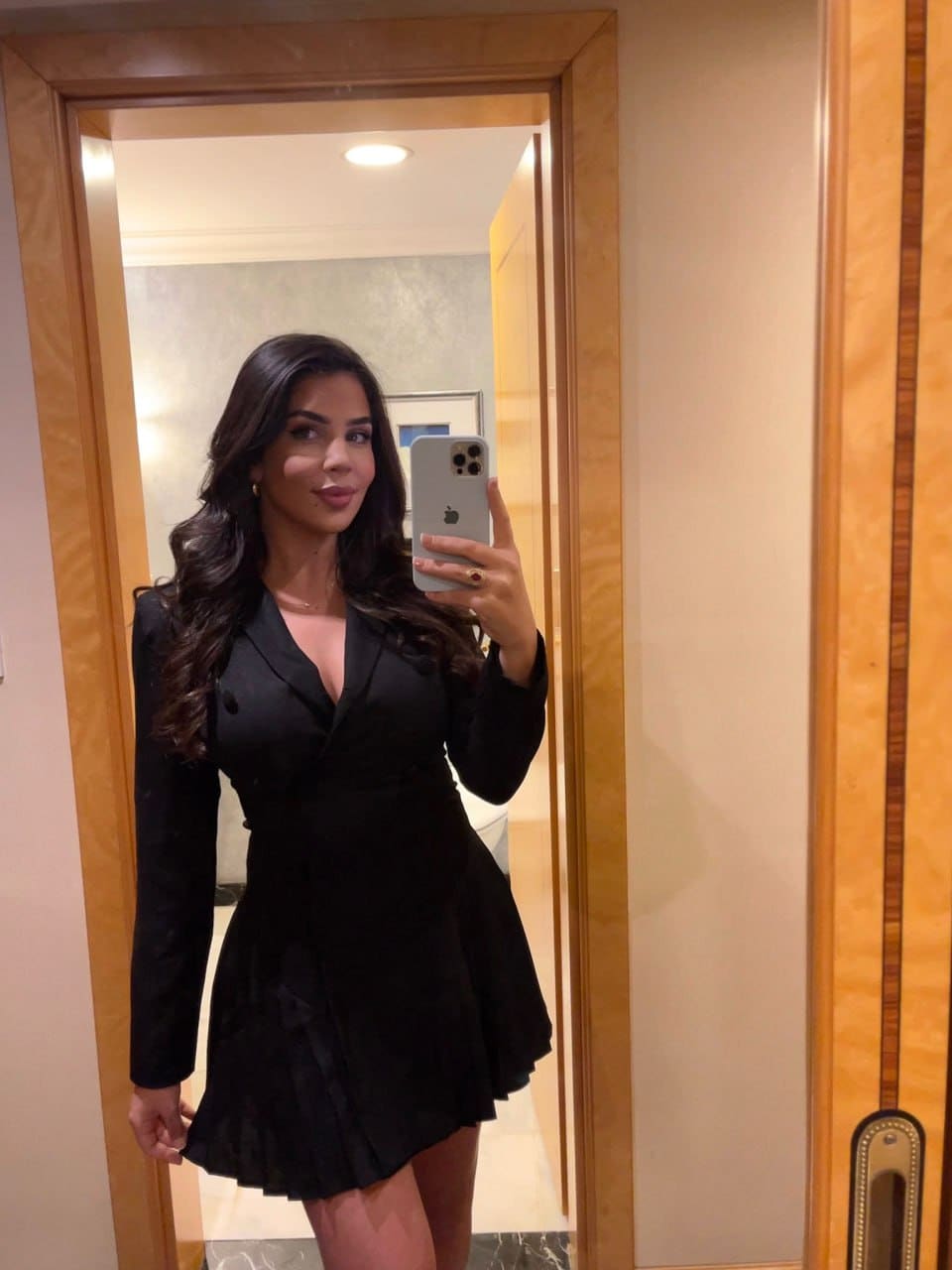 A woman stands before a wooden-framed mirror taking a selfie in a black long-sleeved dress with a deep V-neck and flared skirt.