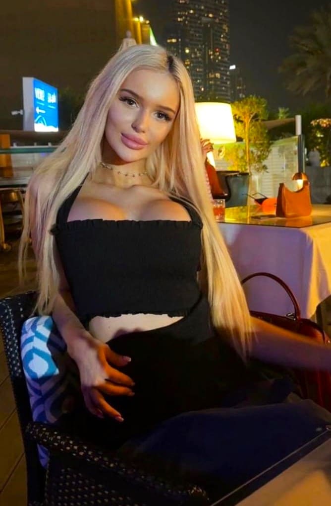 A woman in a black ruffled halter top sits in a woven chair at an outdoor night venue, hand on her midriff.