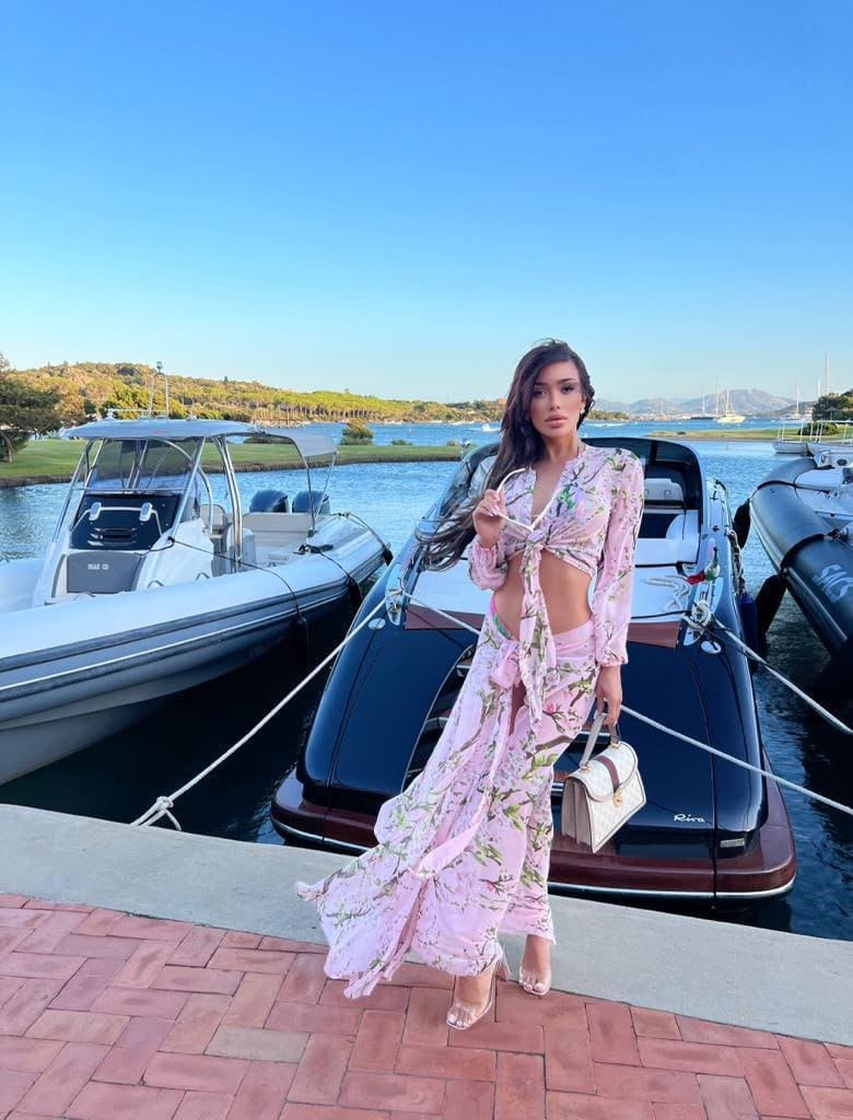 On a brick dock, she wears a pink floral outfit with clear heels and a white bag.